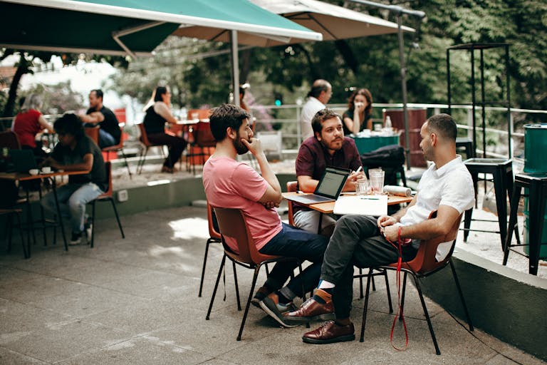 Trois personnes en réunion dans un café en plein air avec des ordinateurs portables et du café. Ils échangent sur le développement d'une application web